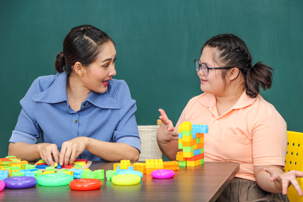 Image of a girl with a disability is practicing the use of hand and finger muscles using blocks and toys and a teacher near her helping her.