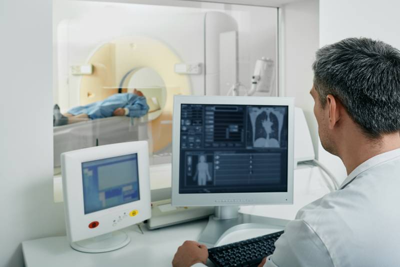 Image of a radiologist viewing the chest CT on his monitor while running a CT scan for a patient lying on the CT scanner
