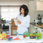 A table full of fruits and vegetables and other healthy food kept inside a kitchen and a woman writing diet plan for herself or a family member.
