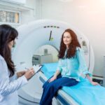 A female doctor holding a writing pad writes down the information given by the patient sitting on the CT table.