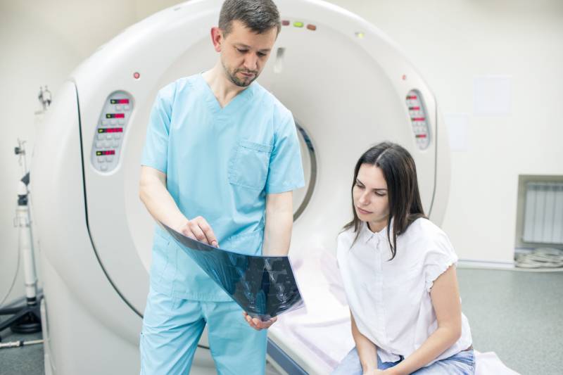 A male technician holding an MRI film explains the scan results to a female patient sitting on the MRI table.