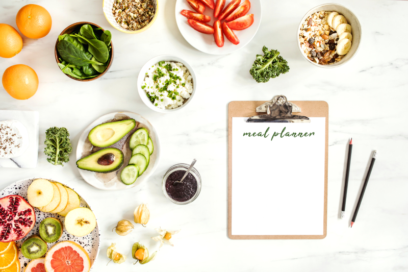 Raw food - organic vegetables, fresh fruits kept in different bowls with a writing pad contatning the word meal planner and 2 pens kept next to it.