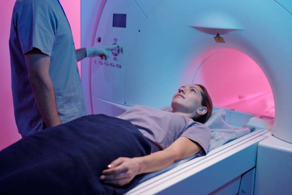 Ayoung woman lying on MRI table and a male technician standing nearby setting the machine for moving her into MRI scanner.
