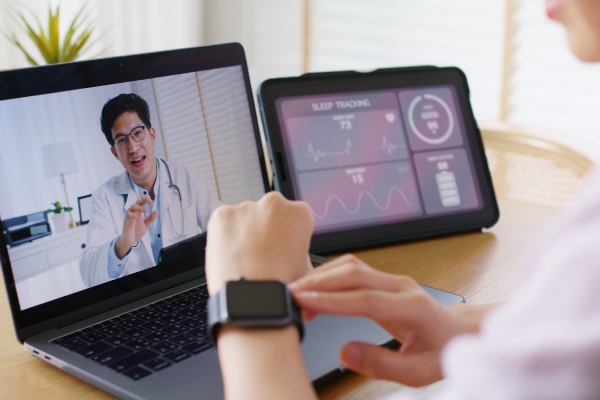Rear side view of a woman checking her smart watch while doing online consultation with a male doctor and a tab kept nearby showing the sleep tracking details on the app.