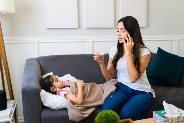 A woman holding a thermometer in one hand and holding a mobile in the other, speaking to someone.Meanwhile her sick daughter is lying on the sofa.
