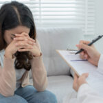 A stressed young woman sitting on a sofa holding her head and a psychiatrist/doctor is recording the patient's condition on a form for treatment purpose.