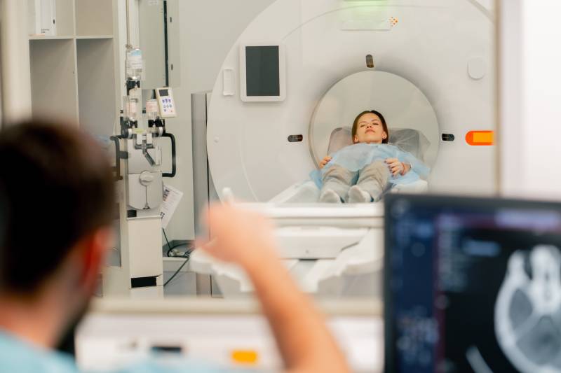 A female radiologist gives instructions to a female patient lying on an MRI scanner.