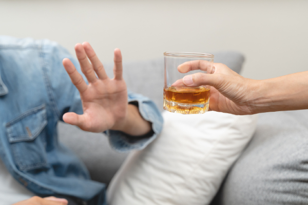 Image of a person's hand offering a glass of alcohol to a man sitting on a sofa and the person raising his hand symbolising "no" or refusing to accept it.