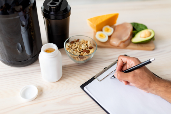 A wooden table with protein rich foods and drinks kept in bowls,bottles,flasks and wooden pieces and a persons hand writing his mean plan on a clipboard kept on the table.