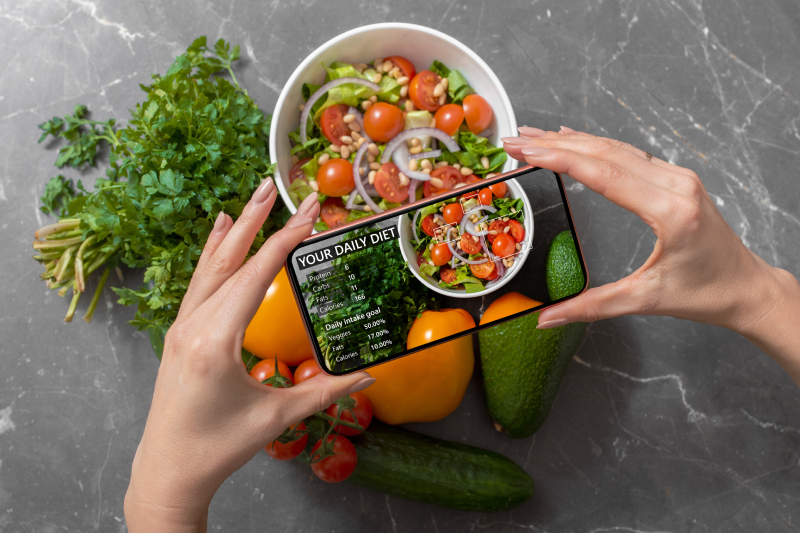 A woman holding a smartphone above food items kept on a table to track nutrition facts and calories in her food.