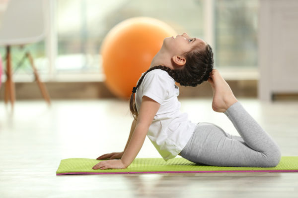 A small girl lying on a yoga mat doing bow pose touches her head with the feet.