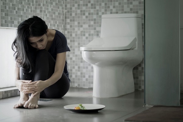 A young woman is sitting behind the western toilet, resting her head on her knee, with a plate beside her and the bathroom door open.
