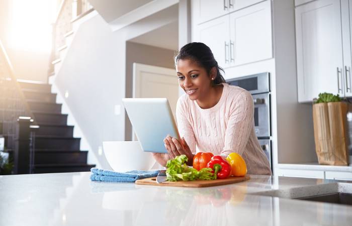 A woman checking her tab sitting on her dining table while preparing to chop vegetables.