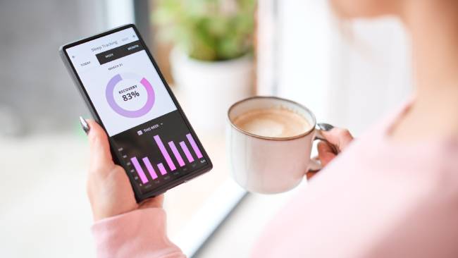 A woman with a cup of coffee looks at her mobile phone in close-up, which shows a sleep tracking app.