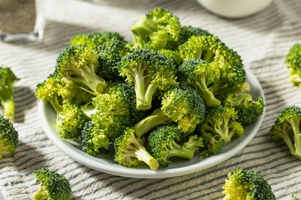 Image of raw cut broccolies kept on a white bowl placed on a white table and few pieces of broccolies scatterd around it.