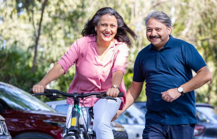 An elderly woman riding bicycle while her husband supports her.