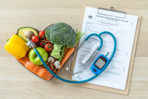 Top view of a table with a stethoscope, a writing pad with a health record chart and a heart shaped bowl containing protein rich foods.