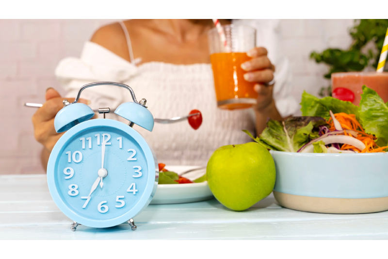 A woman having healthy veg salads, fruits and a cup of juice kept on a dining table along with an alarm showing time as 7:00 AM.
