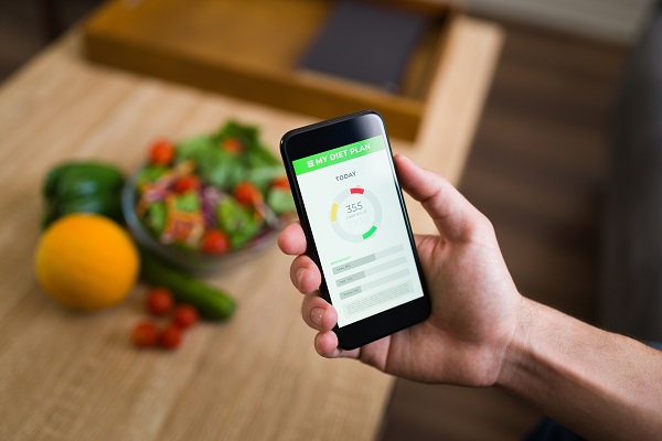 A persons hands holding a smartphone showing the calories of his food in a fitness app and blurred images of fruits and vegetables kept on a wooden table at the background.