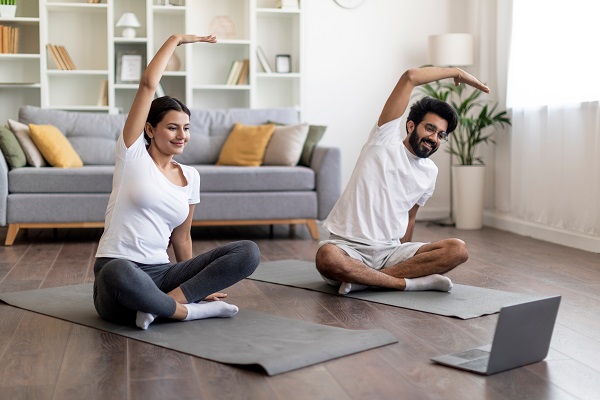 Image of a happy looking couple sitting on yoga mat inside a living room,doing exercise looking at a laptop placed before them.