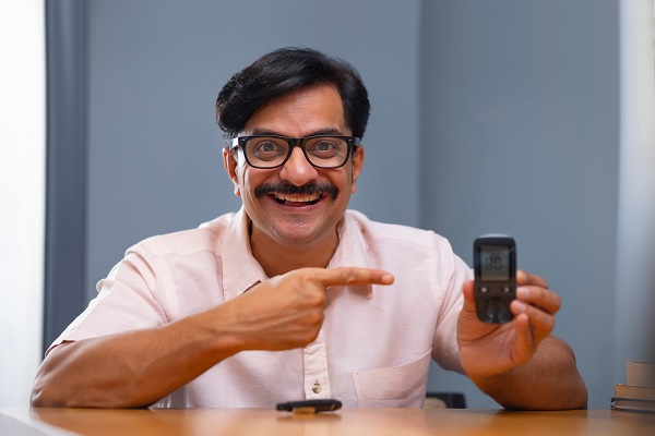 A a man seated at his desk with a happy face,holding a glucometer in his left hand and his right index finger is pointing to the normal sugar level readings on the glucometer.