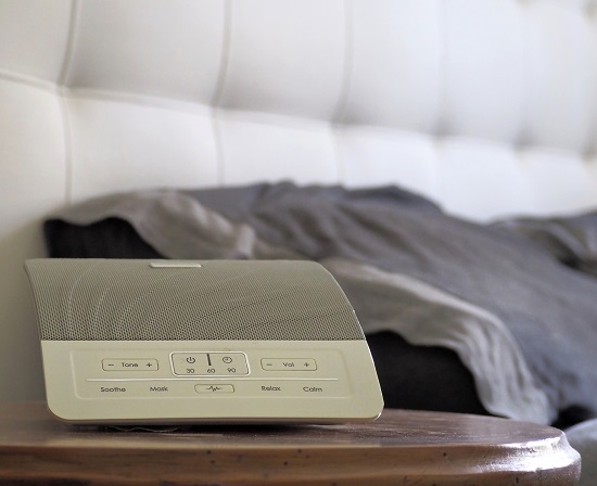 A blurred side view of a bed with a white noise machine placed on the bed side table.