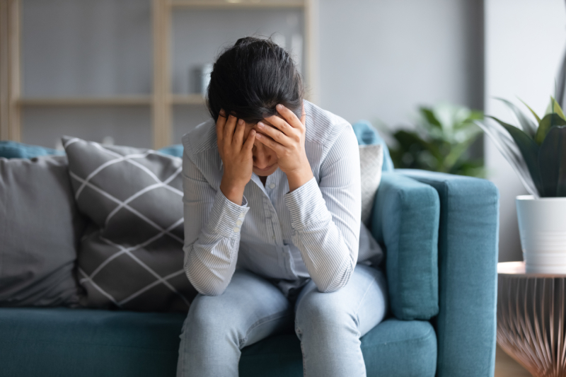 A woman sits alone on the couch, holding her head in her hands, overwhelmed by sadness and loneliness due to worry.