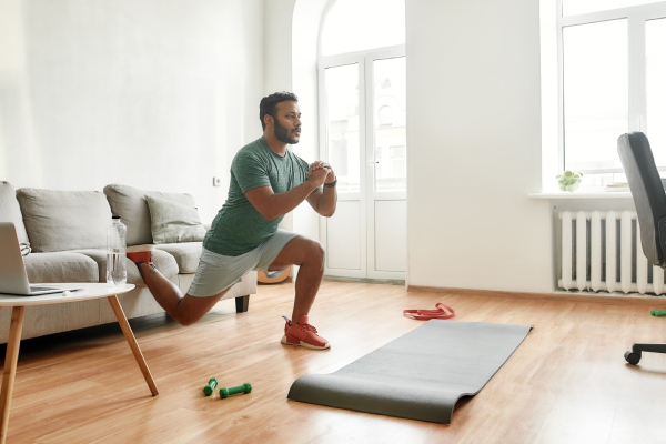 A man performs squat exercises in a bright living room with a yoga mat, dumbbells, and a laptop nearby for an online home fitness session.