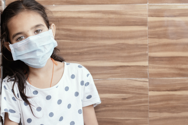 A young girl wearing a medical mask sat, leaning against the wall, overwhelmed by sadness, fear, or anxiety.