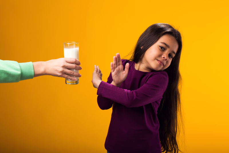 A girl with a dairy allergy is showing a stop gesture in front of a glass of milk on a yellow background.
