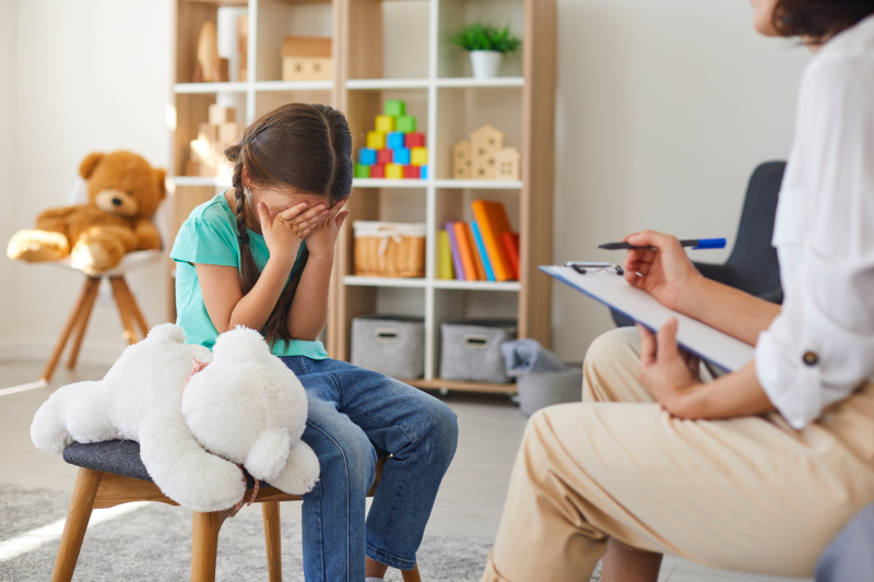 A little schoolgirl is covering her face with emotional stress due to anxiety, fear, or anger, showing behavioral shifts in a psychologist's office.