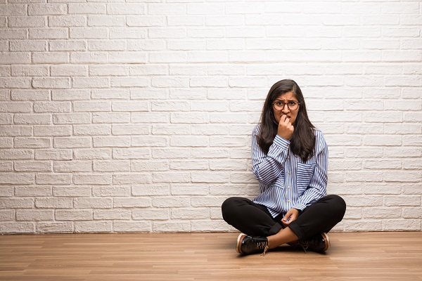 A woman leans against a brick wall, nervously biting her nails, overwhelmed by stress and fear about living a healthy life.