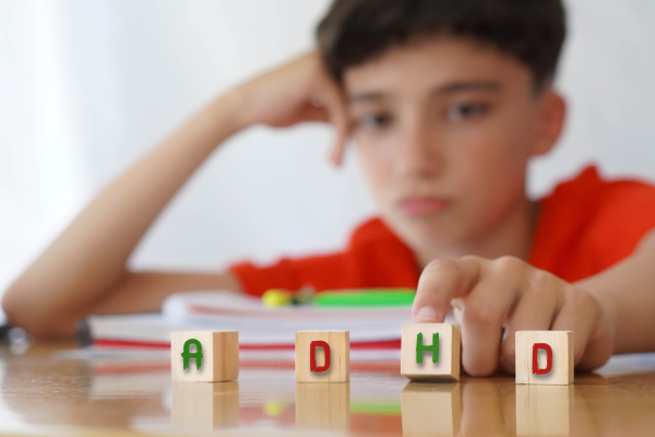 Wooden blocks spelling 'ADHD' are on a table, with a distracted child in the background resting their head on their hand.