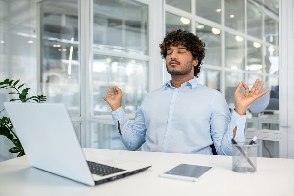 A young man with curly hair sits at his office desk, meditating with eyes closed and hands in a mudra pose, illustrating mental health and stress relief in the workplace.
