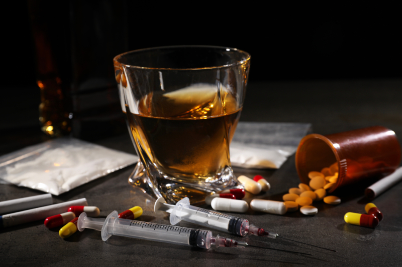 A glass of alcohol, syringes, pills, and cocaine on a table symbolize the significance of tips for overcoming addiction.