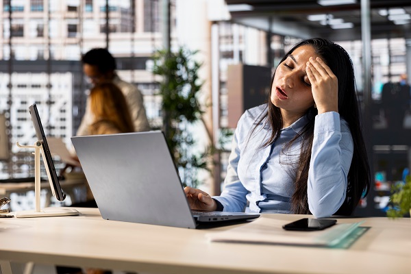 A stressed woman at her desk is showing fatigue, which can be a symptom of iron deficiency and is surrounded by office supplies.