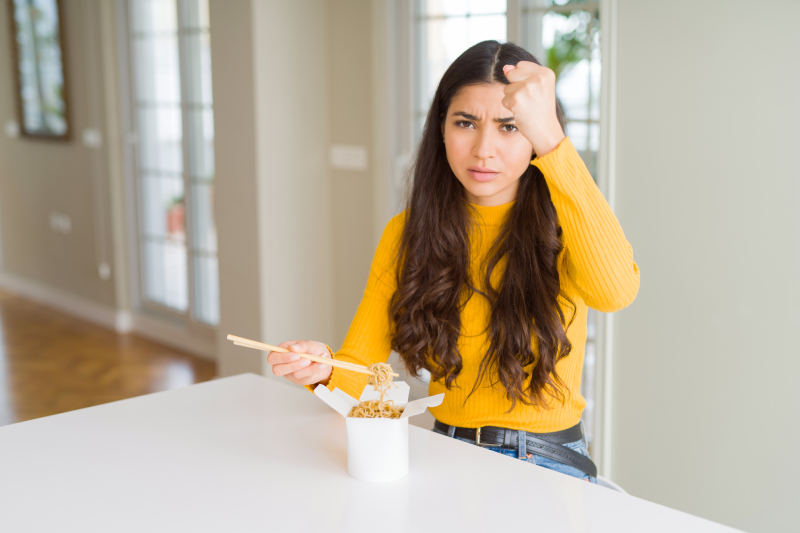 Angry woman eating delivery noodles, with raised hand, highlighting the unhealthy patterns and distress caused by emotional eating.