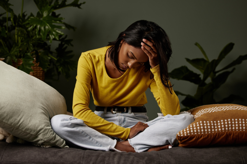 A sad girl holding her head, looking distressed, with pillows and plants around, reflecting the need for mental health tools.