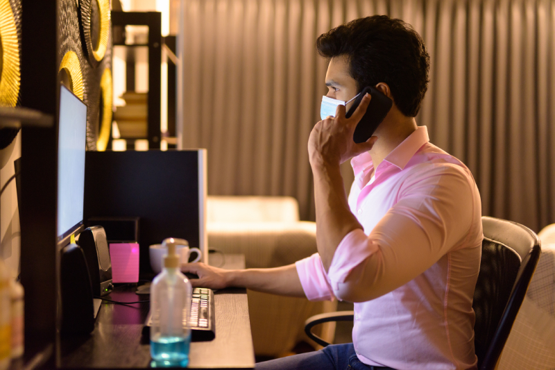 A professional in a mask working remotely, speaking on the phone, with hand sanitizer beside them, balancing work during quarantine.