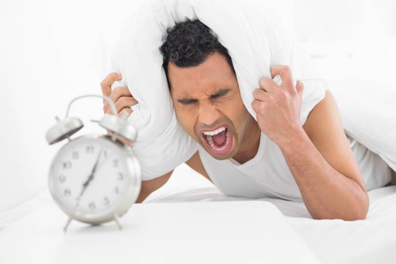 A man woken up by an alarm, covering his ears with a pillow, showing a spike in blood pressure and heart rate.