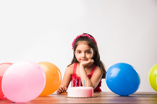 A smiling girl in a red dress sits before a lit birthday cake, surrounded by balloons, symbolizing the challenges children with diabetes face.