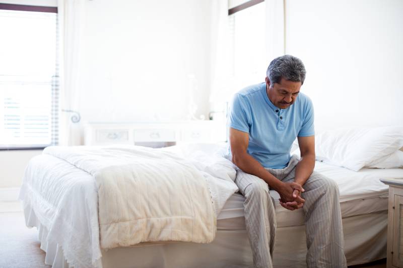 A senior man sitting on a bed, looking down at his hands, representing the link between sleep deprivation, cortisol, and stress.