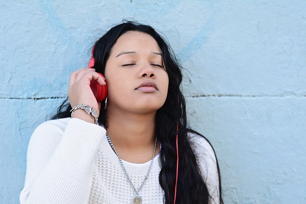 Woman wearing headphones, reclining with eyes closed and hands behind head, symbolizing stress relief by hearing music.
