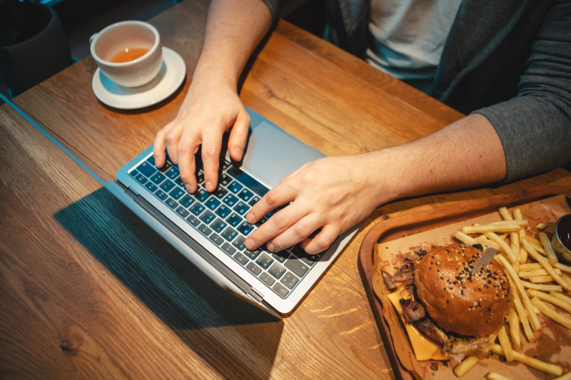 A person at a laptop eating French fries with sauce reflects unhealthy eating habits that can lead to high cholesterol.