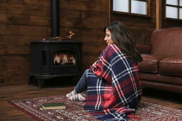 A woman wrapped in a blanket by the fireplace, illustrating the importance of wearing comfortable clothing and maintaining a warm home.