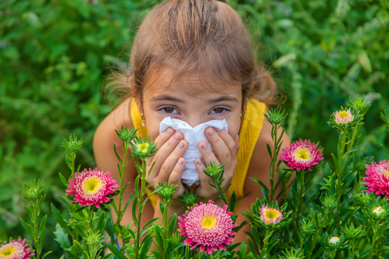 Young girl holding a tissue to her nose in a flower-filled setting, showing signs of a pollen allergy.