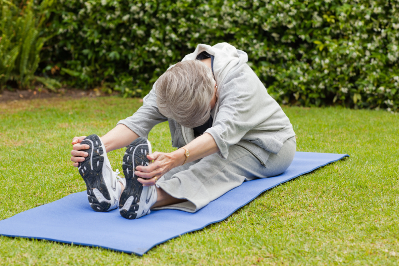 An elderly woman sitting and stretching outdoors, demonstrating proper exercise for arthritis.