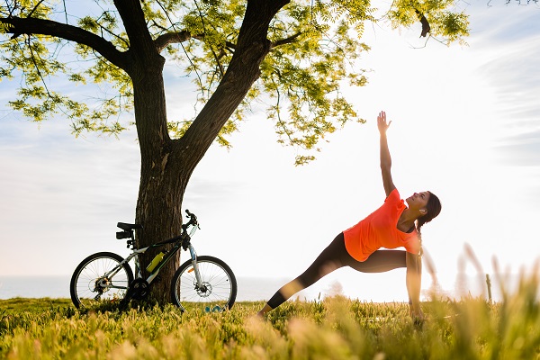 Young woman practicing yoga outdoors near a tree with a bicycle, symbolizing a change in workout environment.
