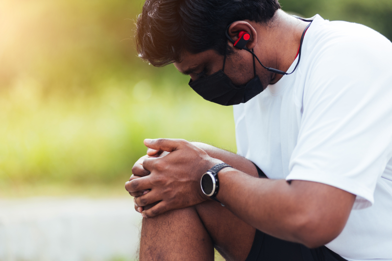 A masked man with headphones holds his knee, symbolizing arthritis and its emotional impact.