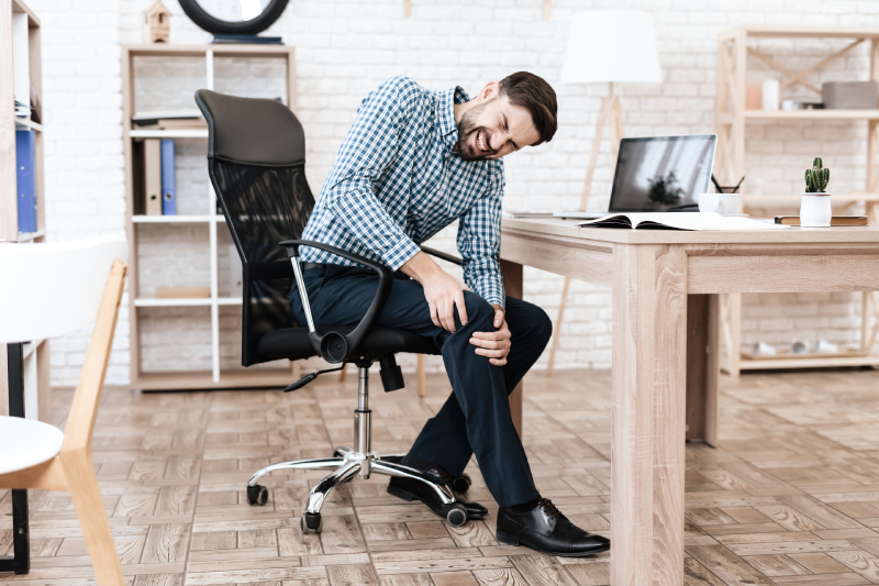 A man sitting in an office chair, holding his painful knee, indicating the need for an ergonomic assessment.